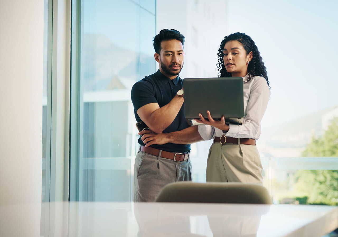 A man and a woman are talking in an office room while standing, with the woman is holding a laptop in her hand.