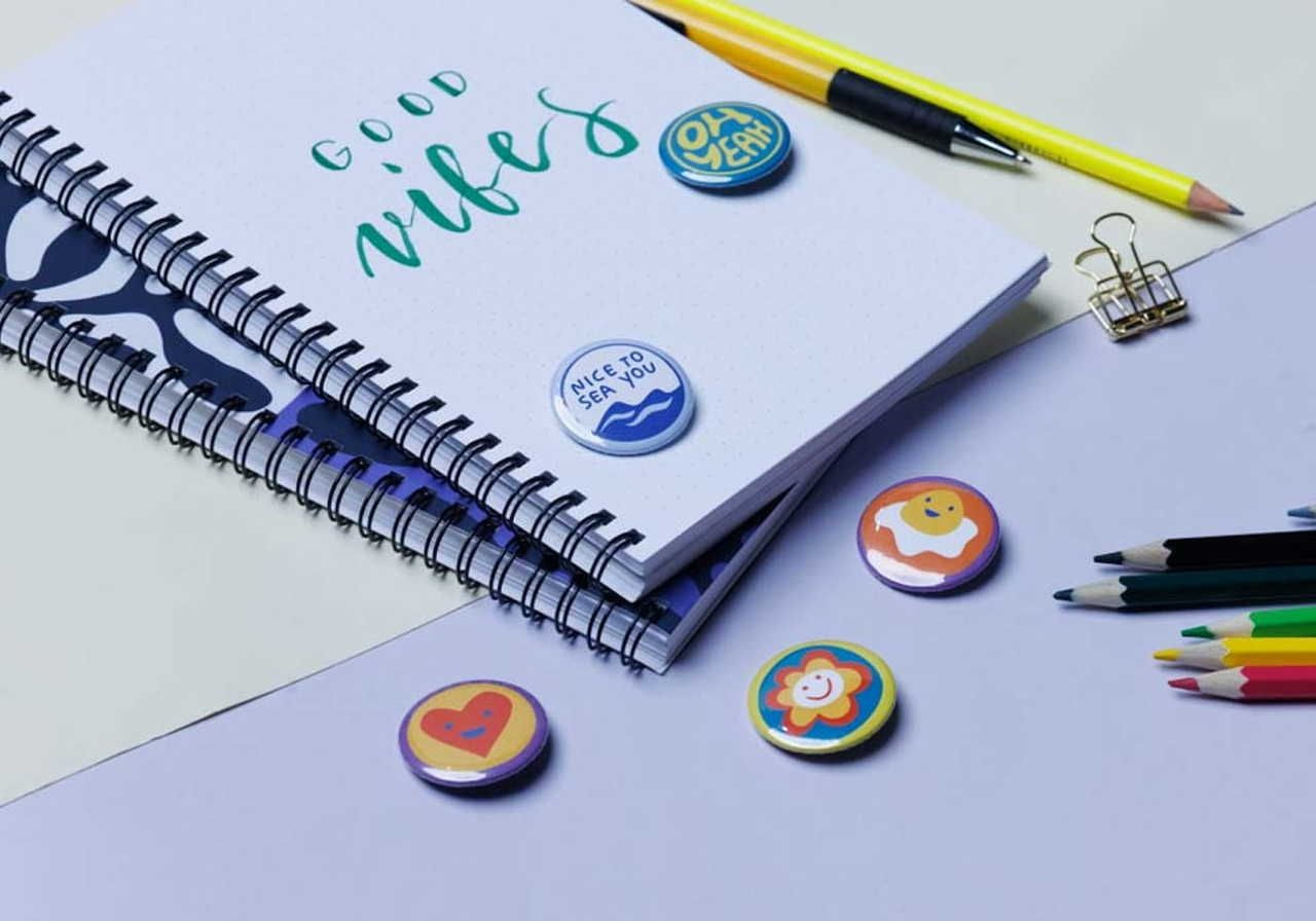 A notebook with the text "Good Vibes," several colorful buttons, a yellow pen, a binder clip, and a selection of colored pencils on a light background.