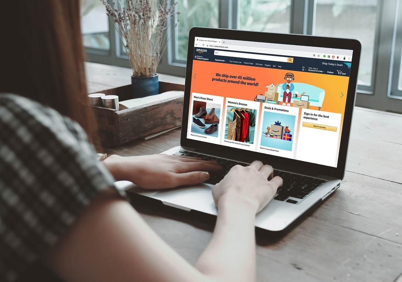 A woman is sitting at a desk with an open laptop and the Amazon homepage on the screen.