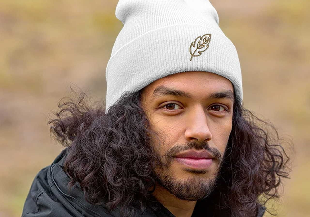 A man with long, curly hair wears a white beanie with a leaf emblem. The background is a soft-focus outdoor setting.