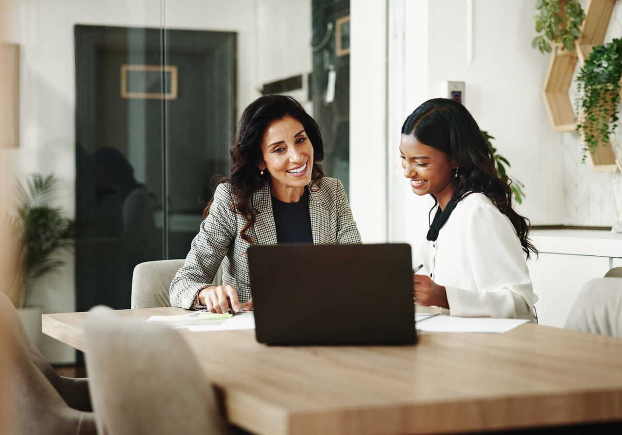 Two women sit at a wooden table in a modern office, smiling while looking at a laptop.