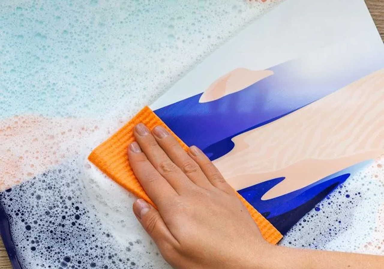 A hand wipes an abstract sunset-themed metal print with a soapy cloth.
