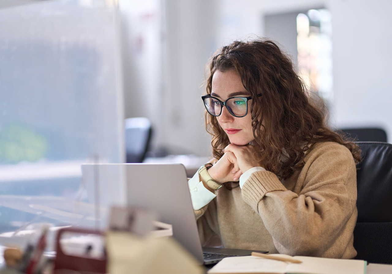 A woman with curly hair and glasses focuses intently on her laptop in an office setting. She wears a beige sweater.
