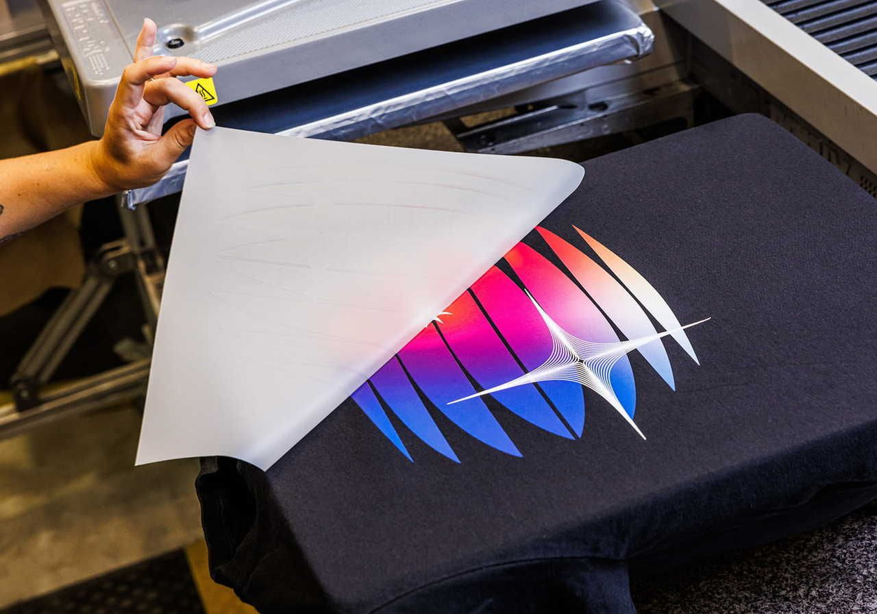 A person holding a translucent sheet above a black t-shirt with a colorful graphic design, ready to be removed after DTFlex printing.