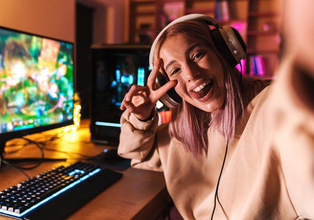 A young woman with headphones smiles, making a peace sign, seated at a gaming setup with a glowing keyboard and monitor.