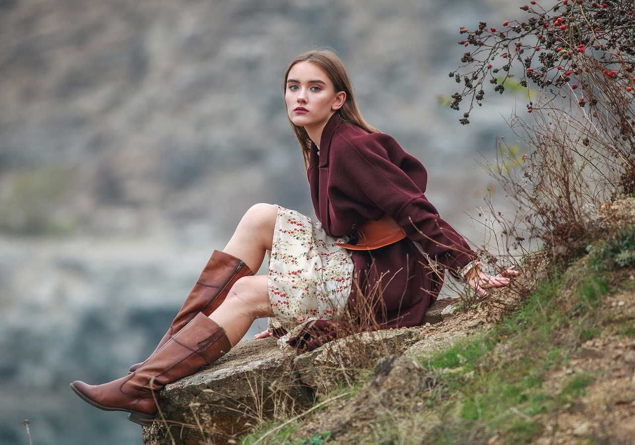 A woman in a maroon coat and floral dress sits on a rocky ledge, surrounded by dry grasses and red berries.