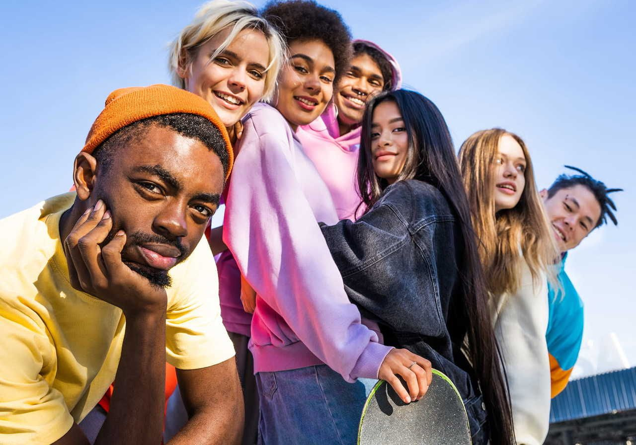 A diverse group of young adults smiling under a clear blue sky, wearing colorful casual clothing. One holds a skateboard.