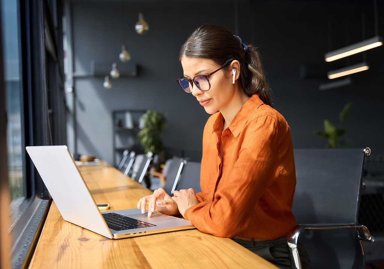 A woman in an orange shirt works on a laptop, searching for dropshipping marketing tips. She wears glasses and earbuds.