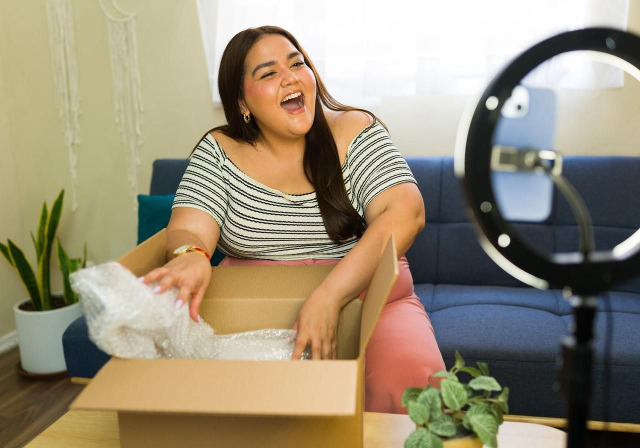 A young woman is filming herself with a mobile phone while unpacking a delivery box.