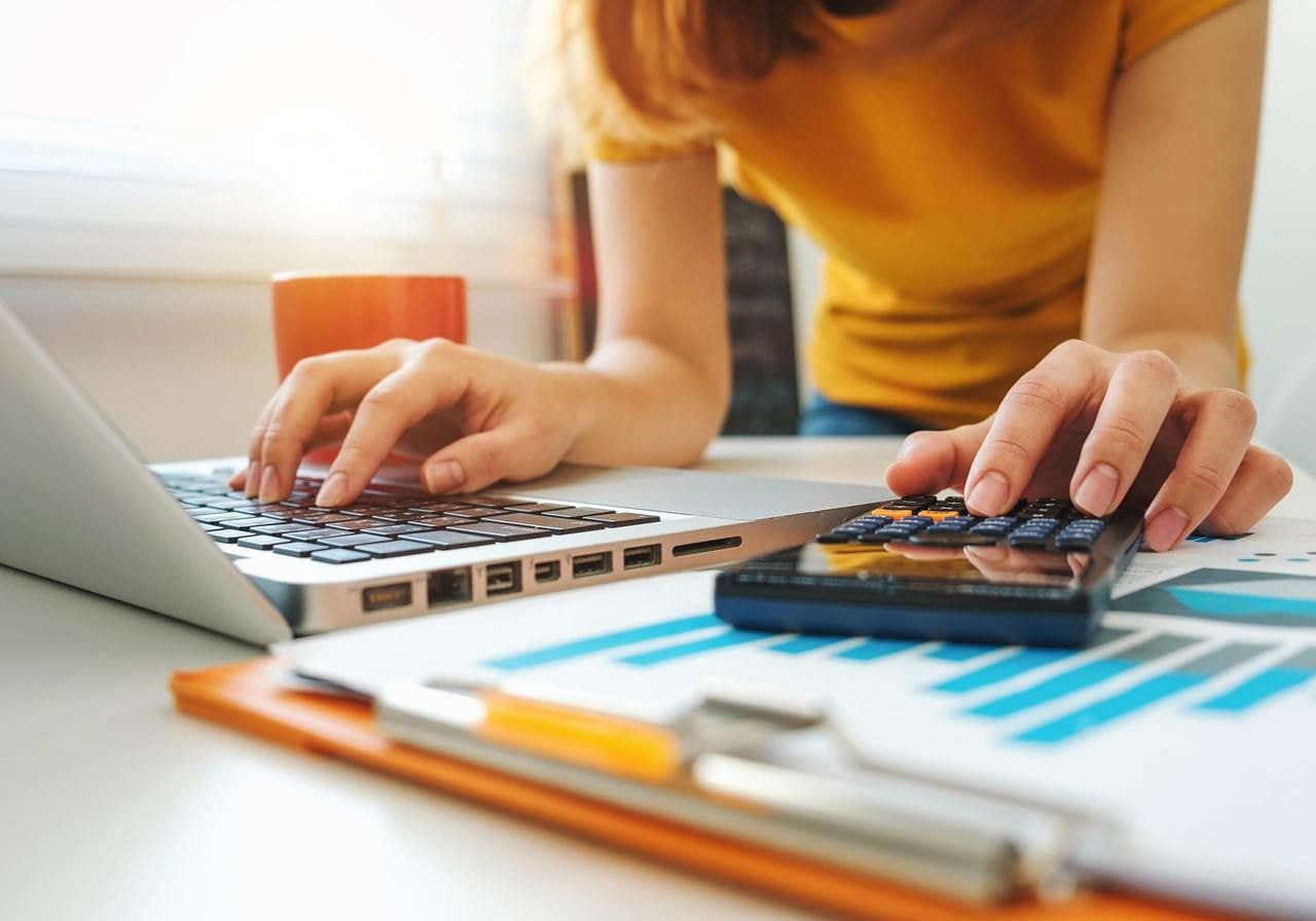 A woman in a yellow shirt uses a laptop and a calculator at a bright desk. Nearby are charts, a clipboard, and a mug.