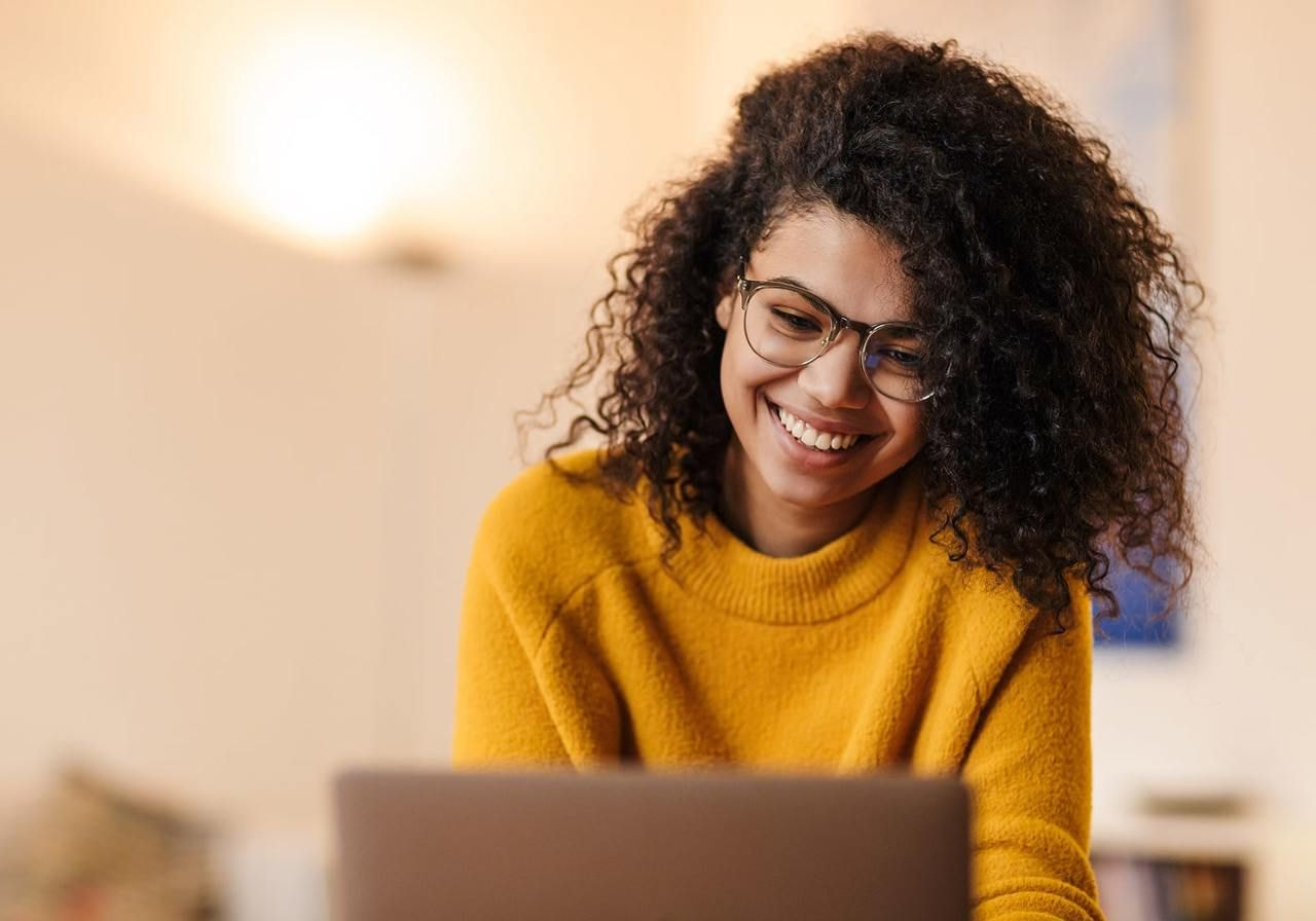 A smiling woman with curly hair and glasses in a cozy yellow sweater, looking at a laptop, searching for a clothing line business plan.