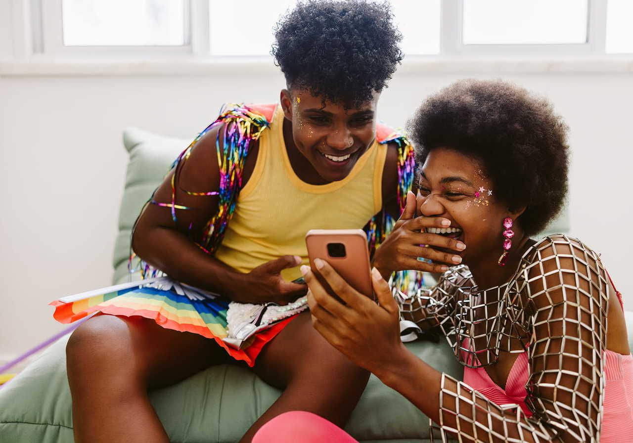 Two friends laughing together while looking at a smartphone, wearing colorful outfits and accessories in a bright room.