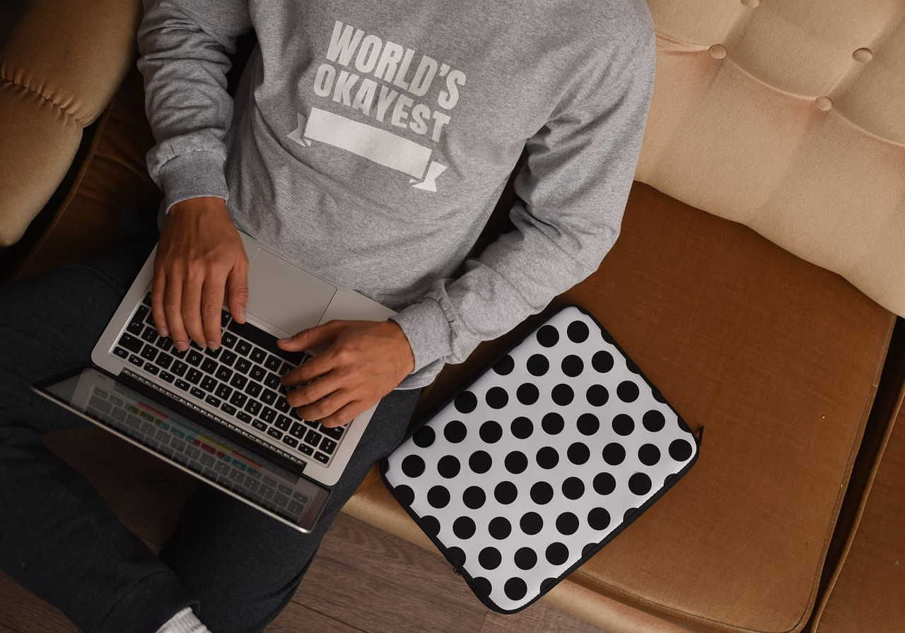 A person wearing a gray sweatshirt sits on a beige couch, using a laptop. There is a black-and-white polka-dot laptop sleeve nearby.