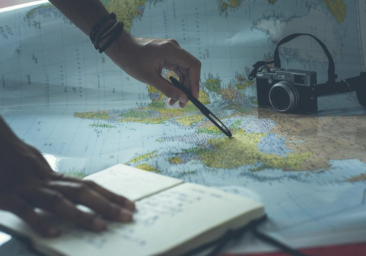 A hand holding a pen and a camera rests on a map, suggesting planning or documenting a journey.