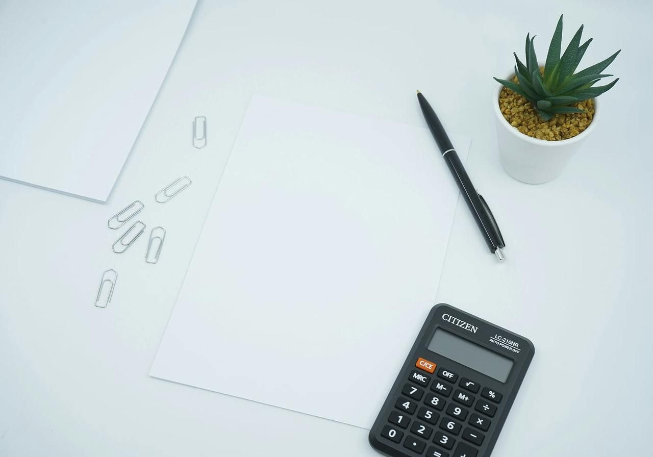 Blank paper, a calculator, a pen, and a small plant arranged on a white table surface.