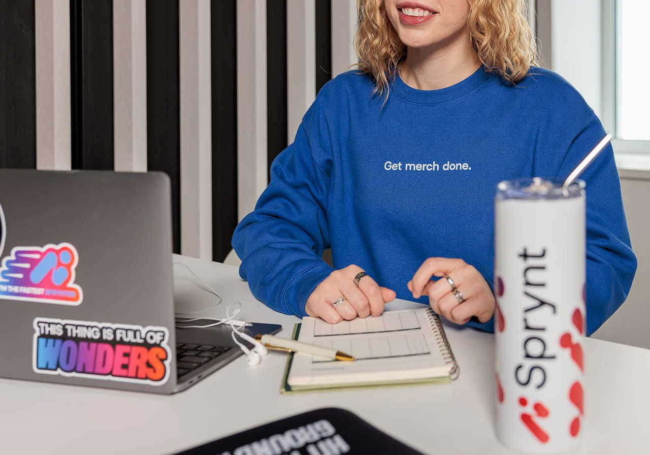 A woman with curly hair wearing a blue sweatshirt sits at a table with a laptop, a notepad, and a drink.