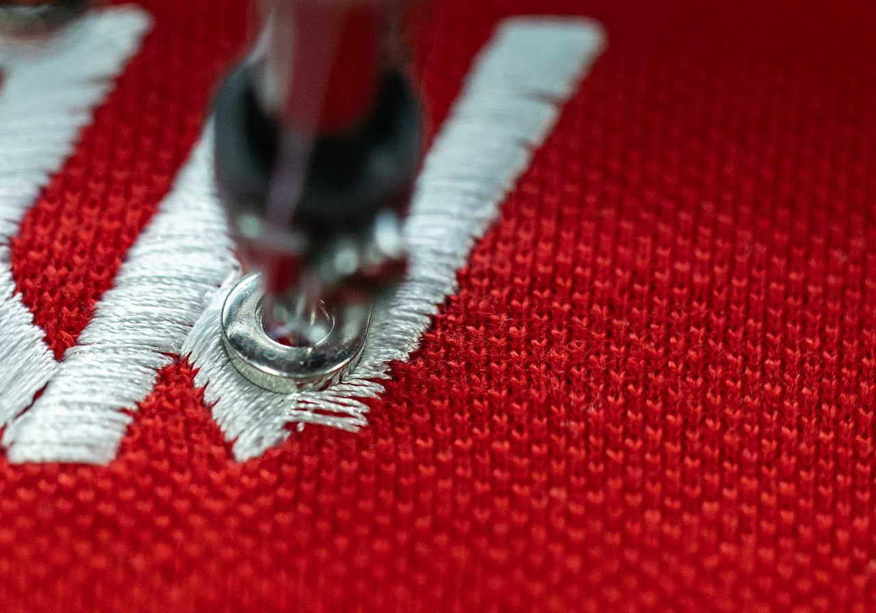 Close-up of a sewing machine embroidering white thread on red fabric, creating a "W" shape.