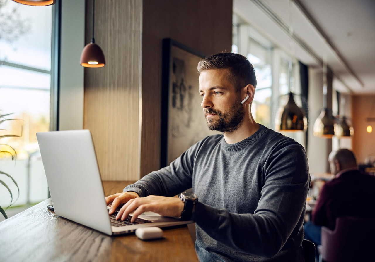 A bearded man wearing earbuds works on a laptop at a table in a cafe.