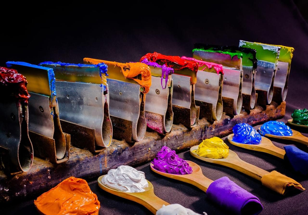 A row of paint scrapers covered in vibrant red, blue, orange, pink and green paints sits above wooden spoons.