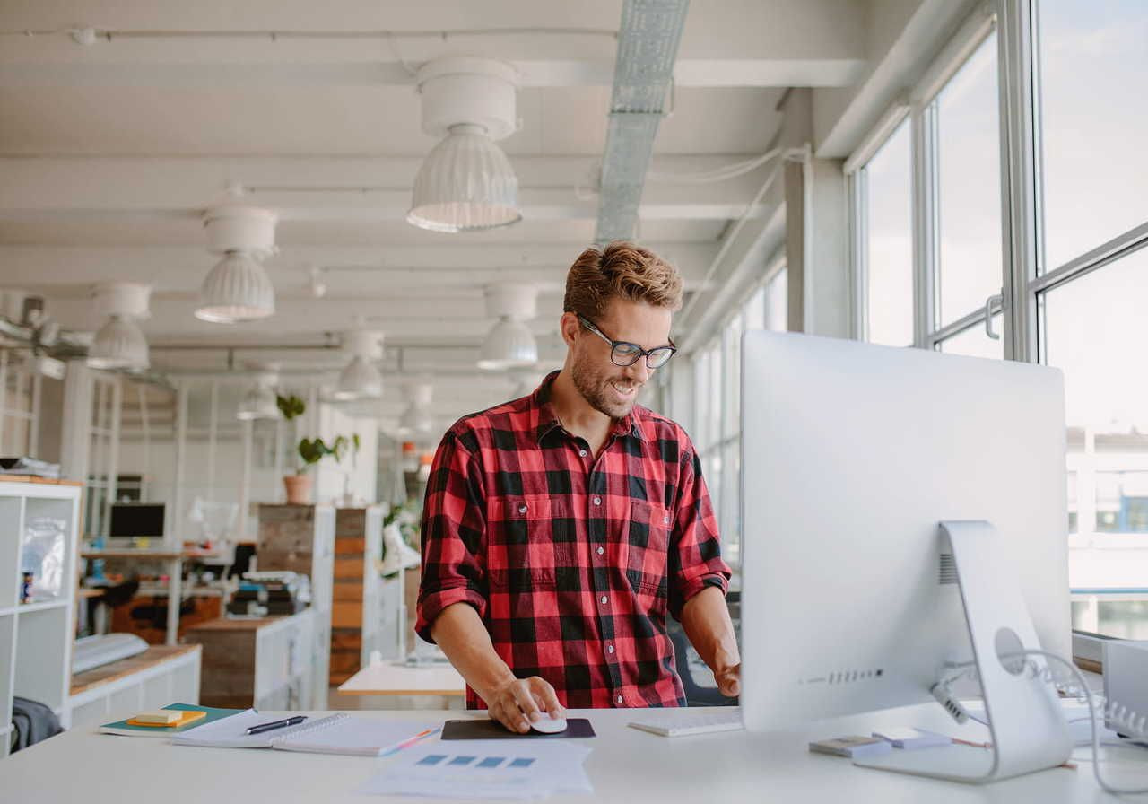 A smiling man is doing graphic design work at a standing desk, with a monitor, notebook and papers on top of it.