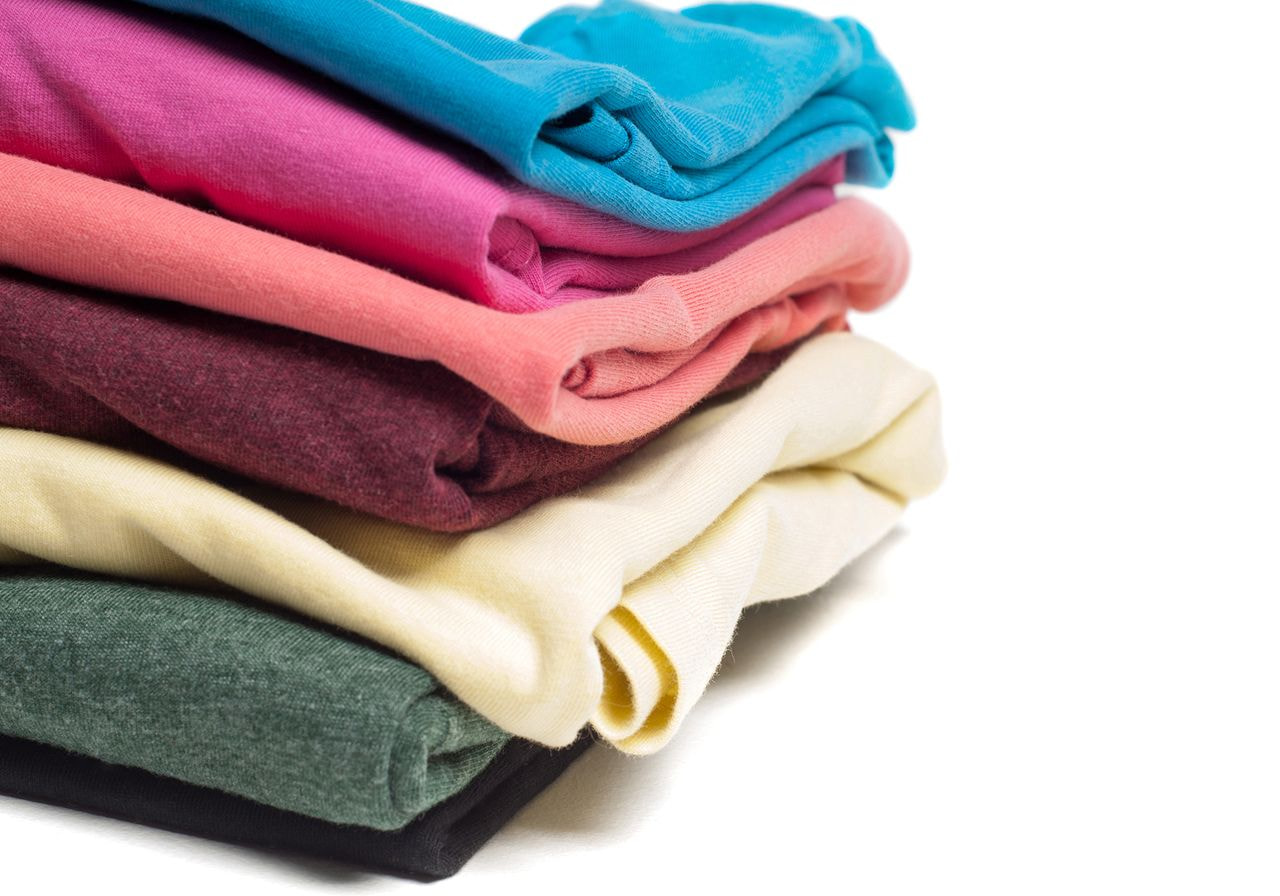 A stack of folded blank t-shirts in blue, pink, coral, maroon, cream, green, and black sits on a white background.