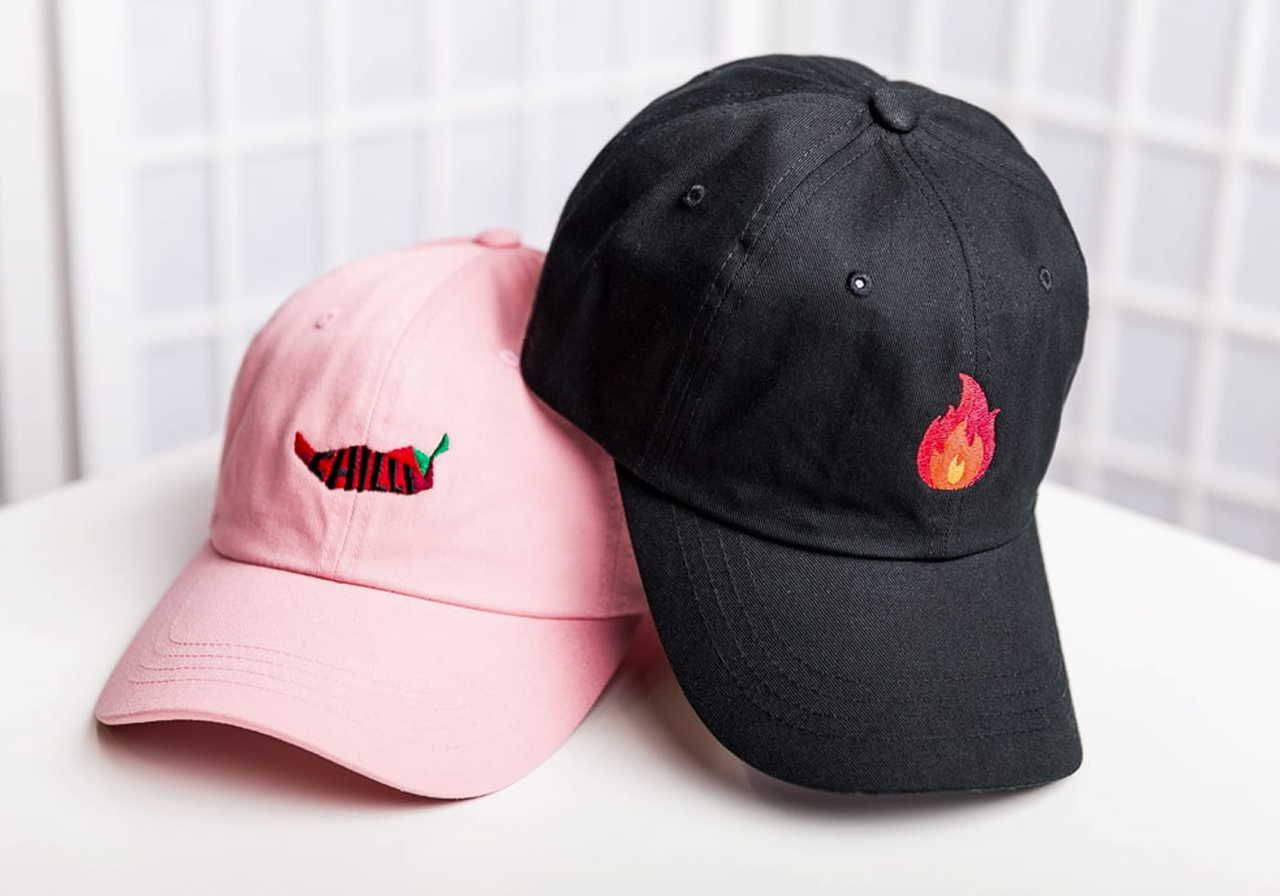 A pink baseball cap with a chili pepper design and a black baseball cap with a flame design.