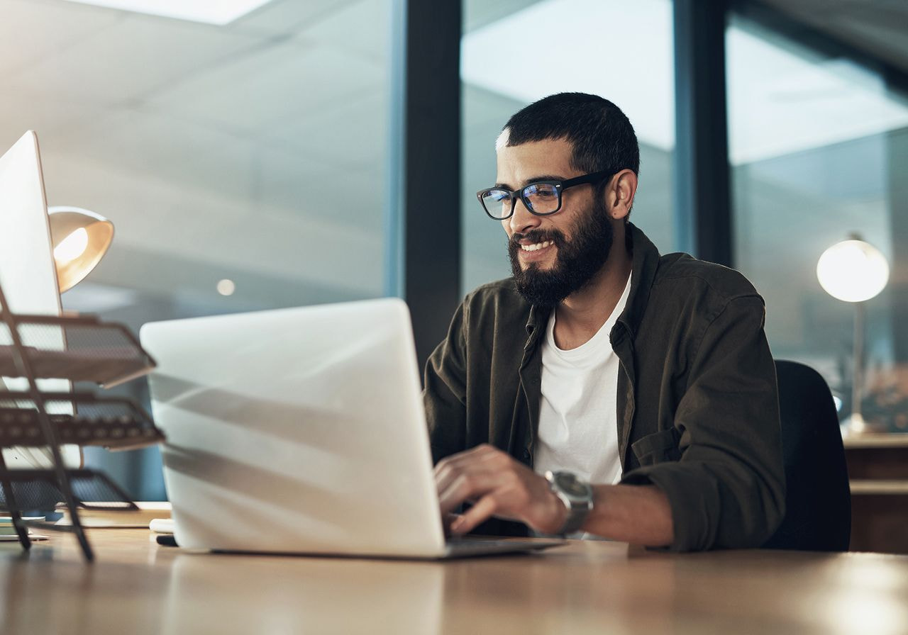 A smiling man in glasses types on a laptop at his desk in a dimly lit office.