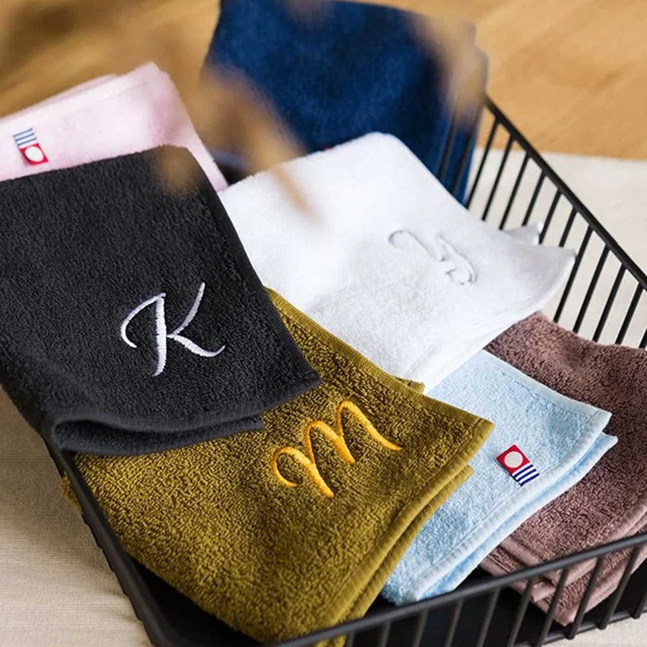 A stack of colorful towels with embroidered letters in a basket. Towels in black, white, mustard, brown, and blue.
