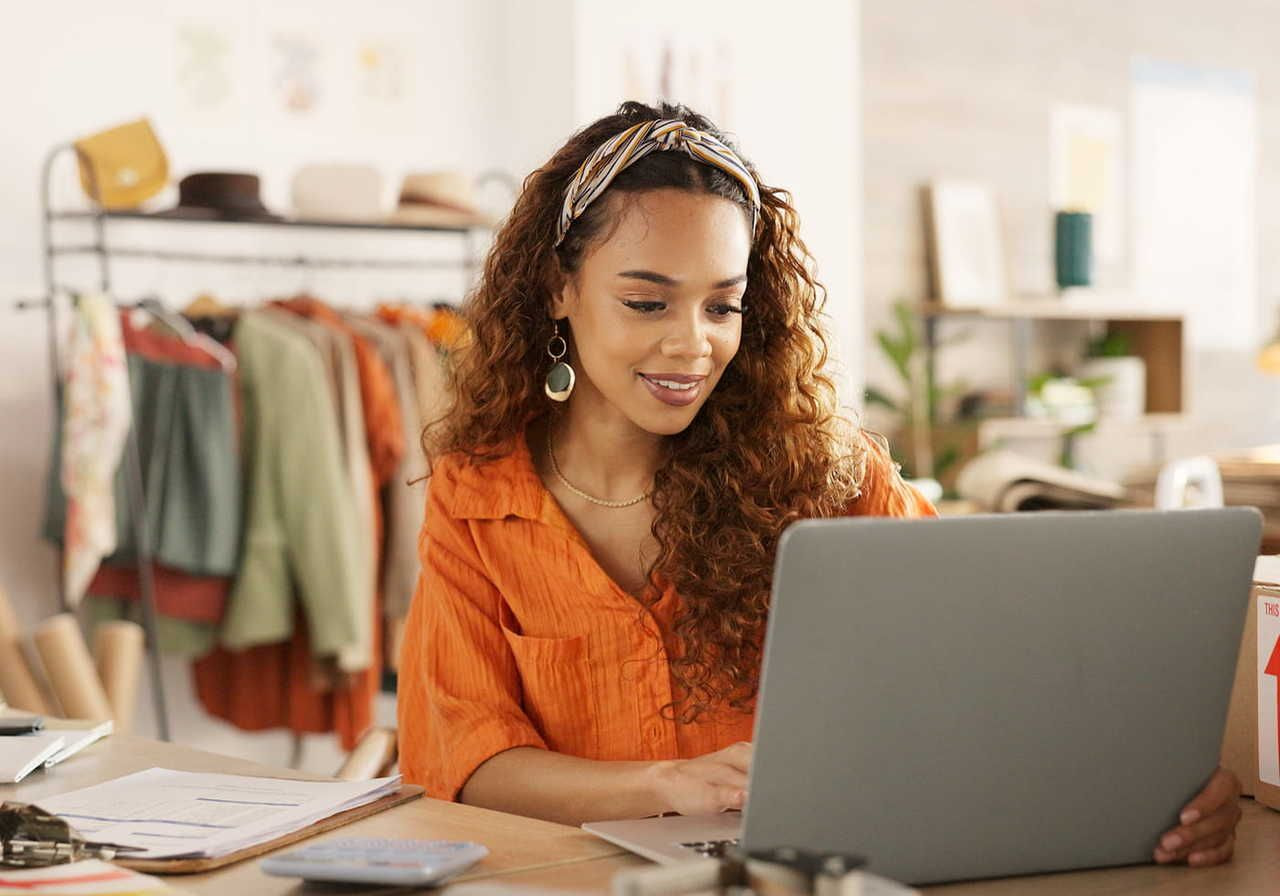 A woman in an orange shirt works on a laptop, comparing DTG vs embroidery, in a cozy room with racks of clothes in the background.