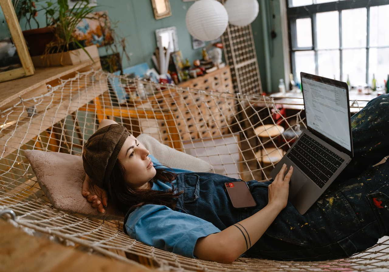 A woman in a cap and blue overalls relaxes on a hammock indoors, using a laptop.