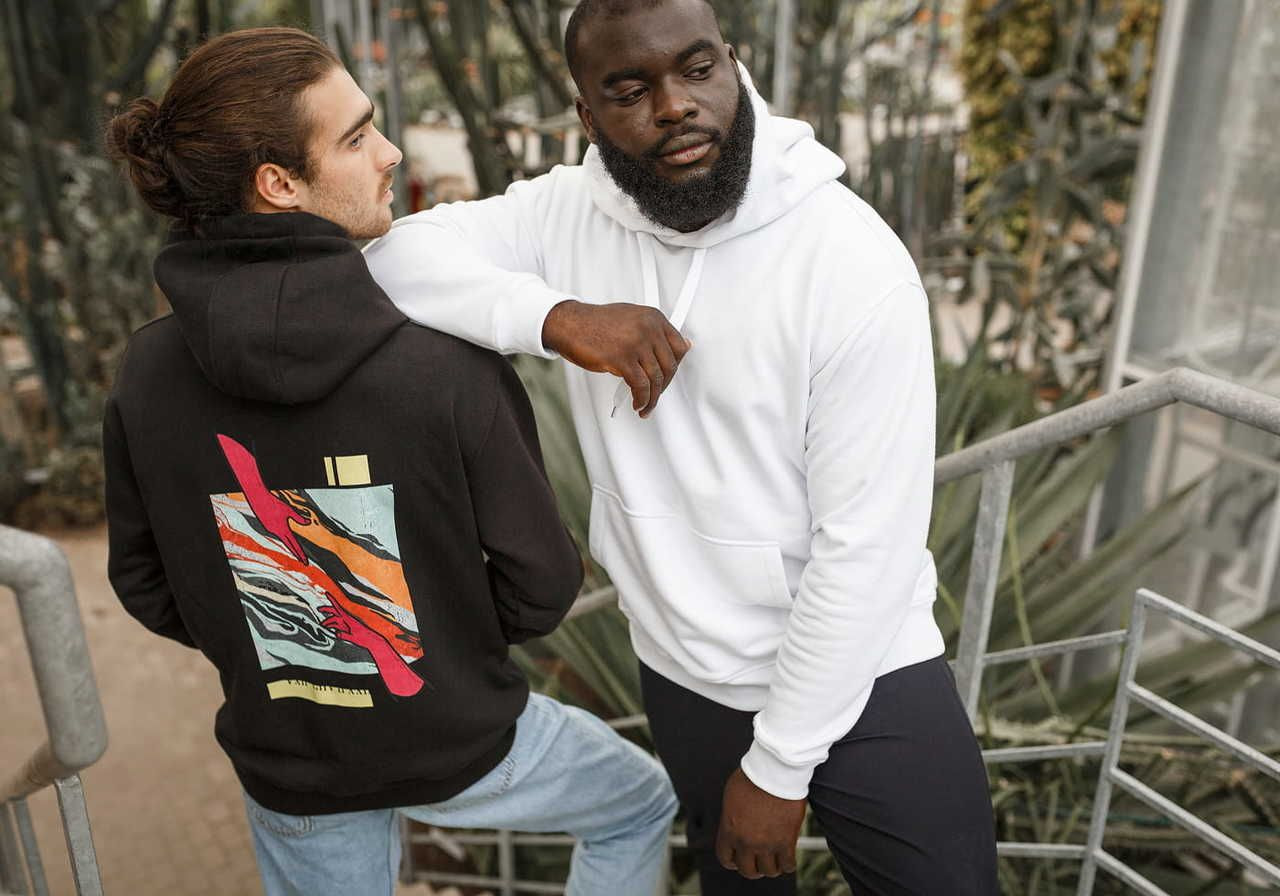 Two men are standing on a staircase, one wearing a black hoodie with a colorful design on the back and the other in a white hoodie.