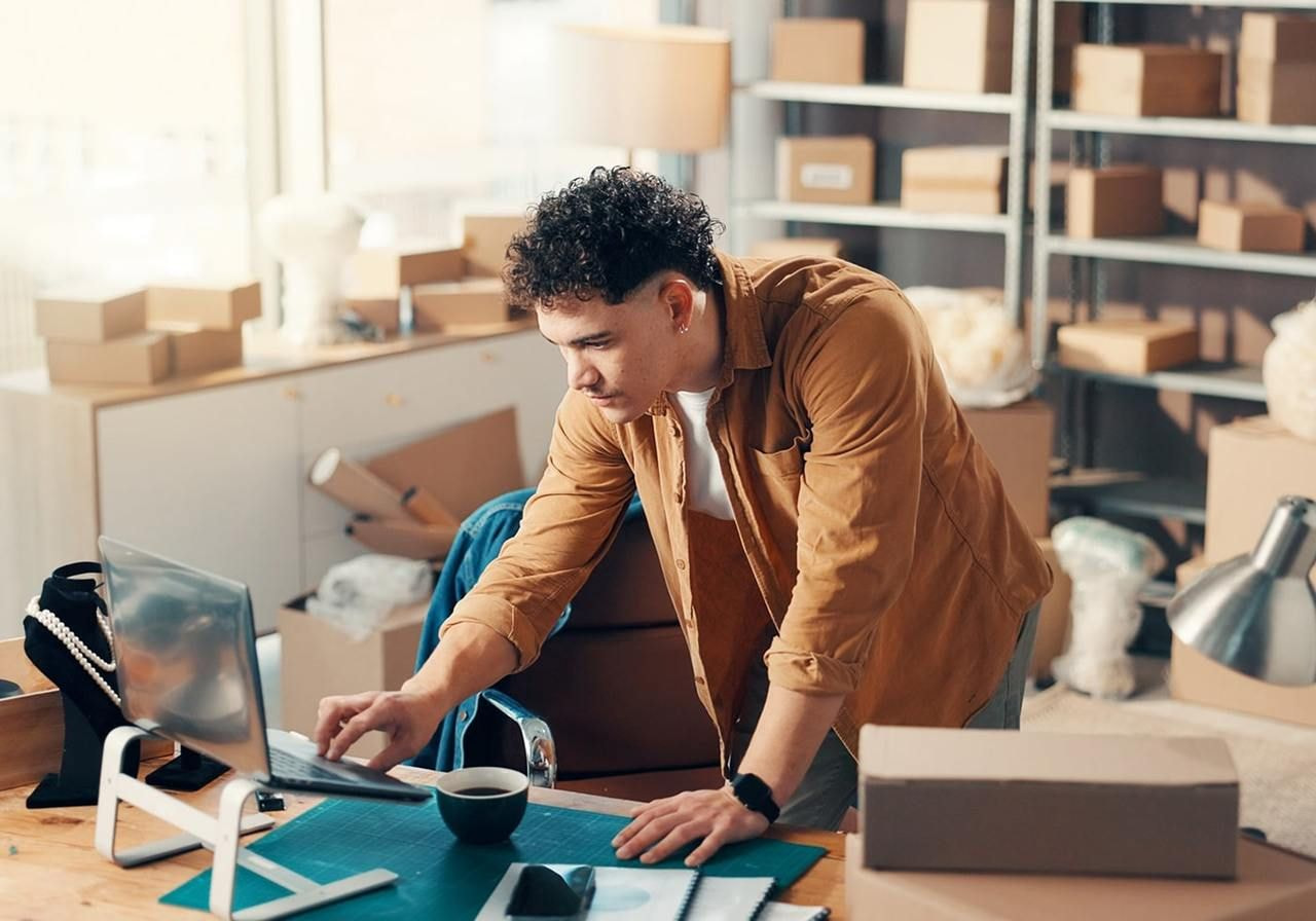 A focused man leans over a laptop, learning how to make phone cases, in a home office filled with boxes. He wears a brown shirt.