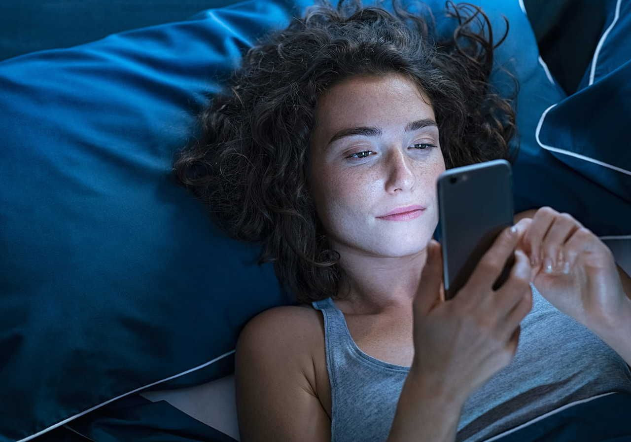 A young woman with curly hair is lying on a bed surrounded by blue pillows, looking at her smartphone with a relaxed expression.