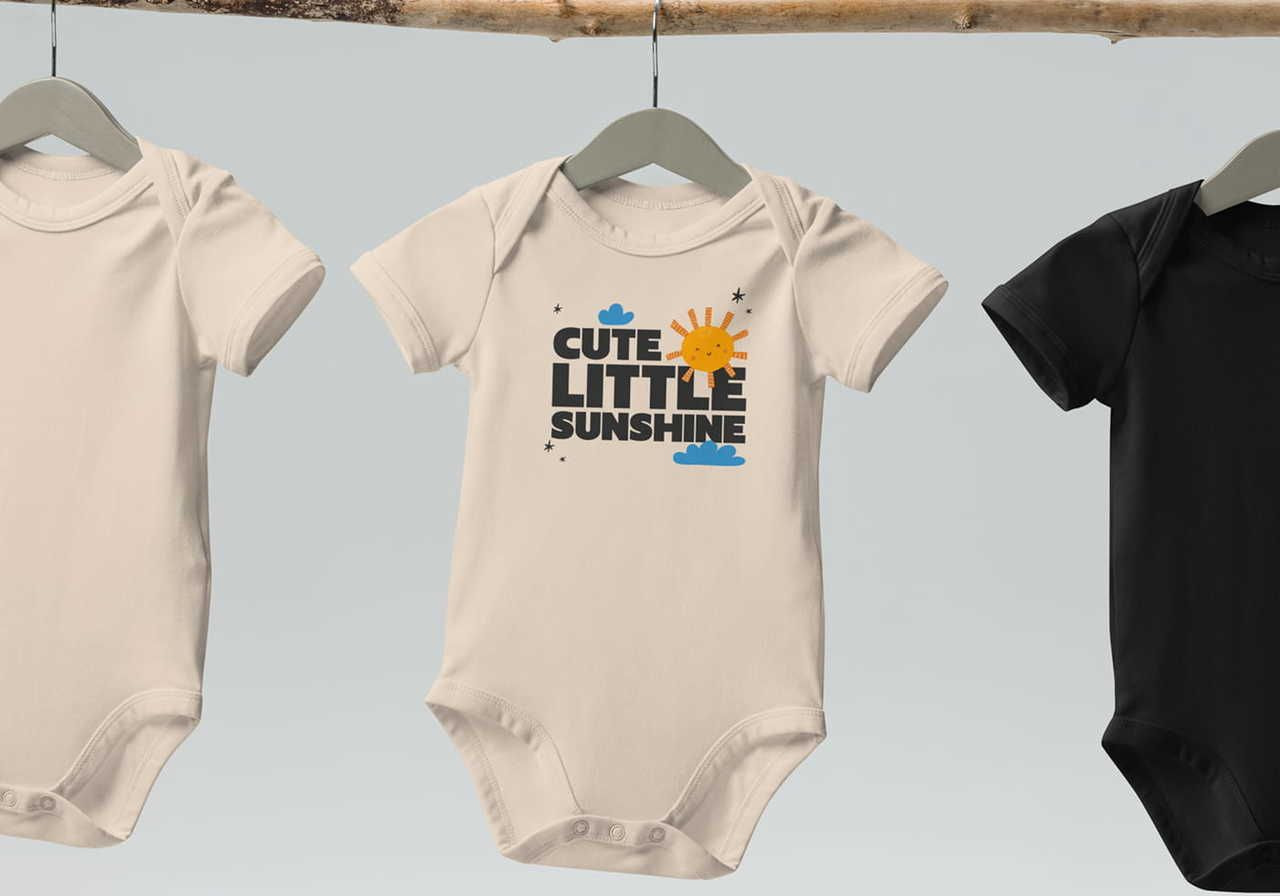 Three baby onesies hang on a wooden rod. The middle one has "Cute Little Sunshine" with a sun graphic. Colors: beige, and black.