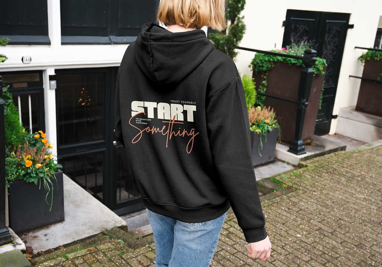 A person in a black hoodie with "Start Something" written on the back walks down a cobblestone street. Flower pots nearby.