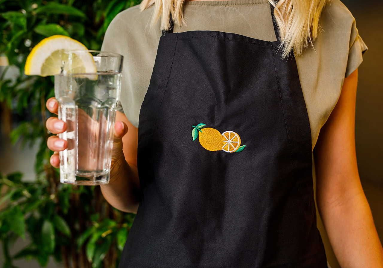 A person wearing a black apron with an embroidered lemon design holds a glass of water with a lemon slice.