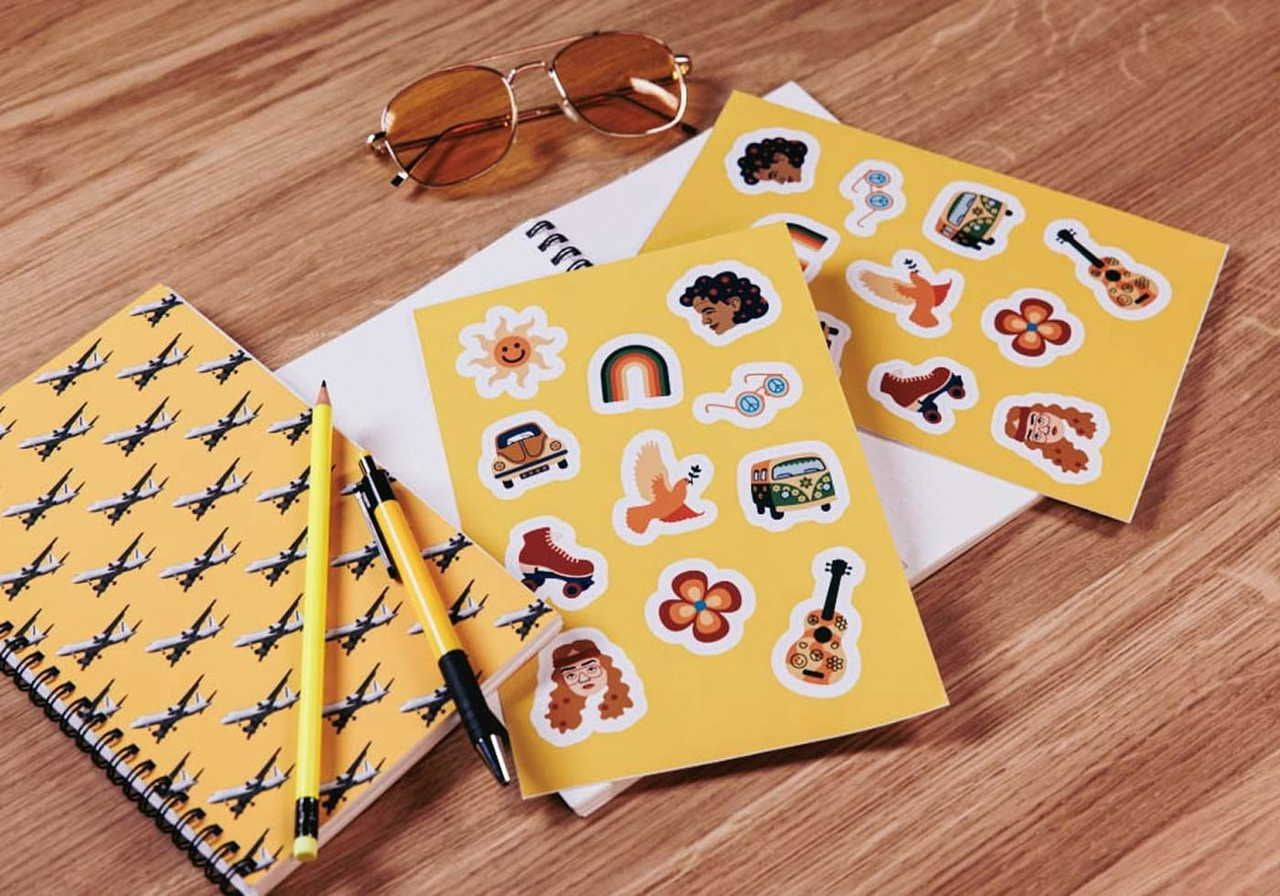 A flat lay of two notebooks and sticker sheets on a wooden surface, with yellow sunglasses and pens.