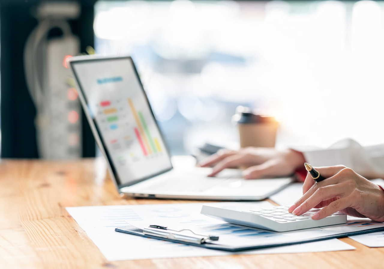 Hands typing on a calculator and a laptop displaying colorful charts on a desk, papers, and a coffee cup nearby.