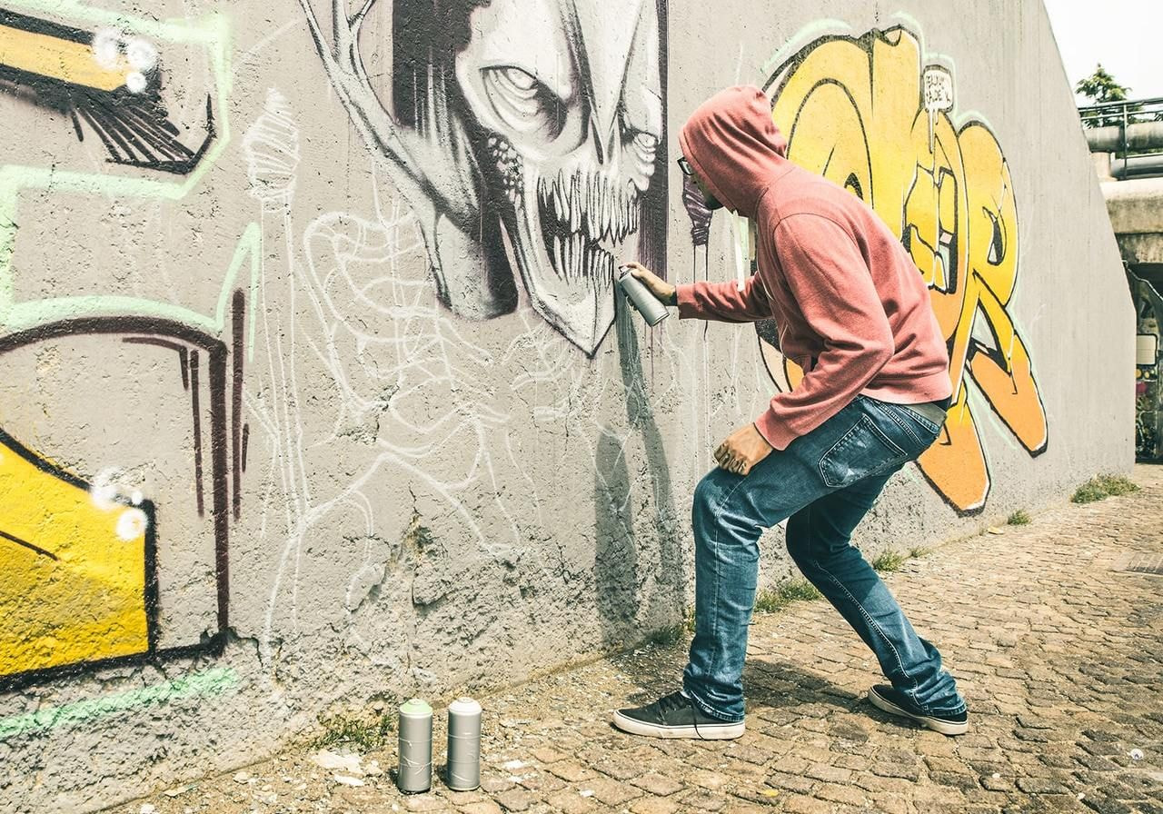 A person in a red hoodie spray-paints a large, detailed monster face on a graffiti-covered wall.