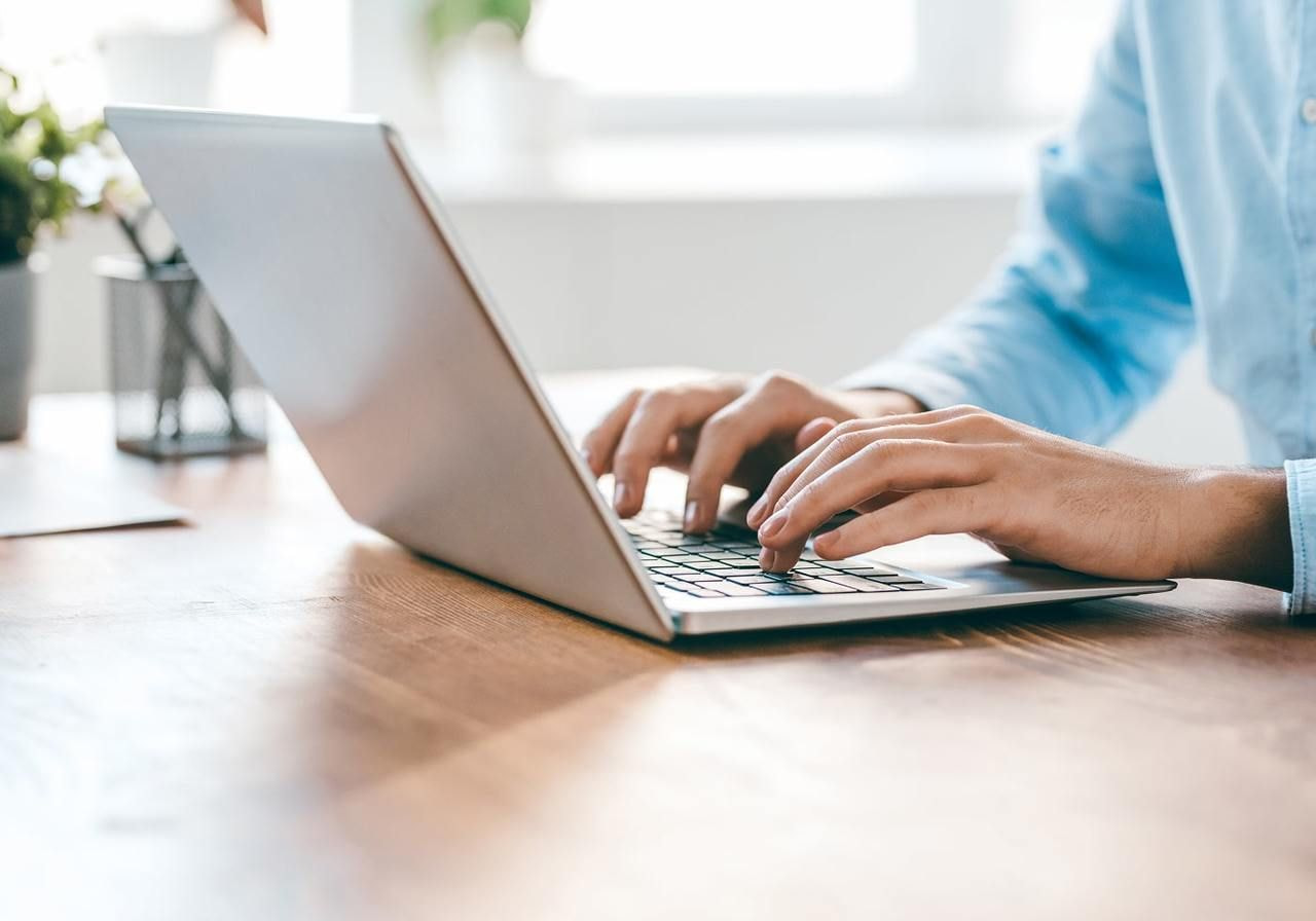 A man’s hands typing on a laptop on a wooden desk. A small plant and pen holder are in the background.