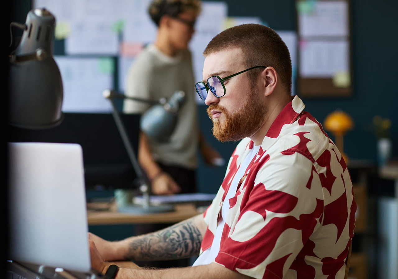A man with glasses and a red-patterned shirt works on a laptop, searching for how to sell mugs on Etsy, in a modern office.