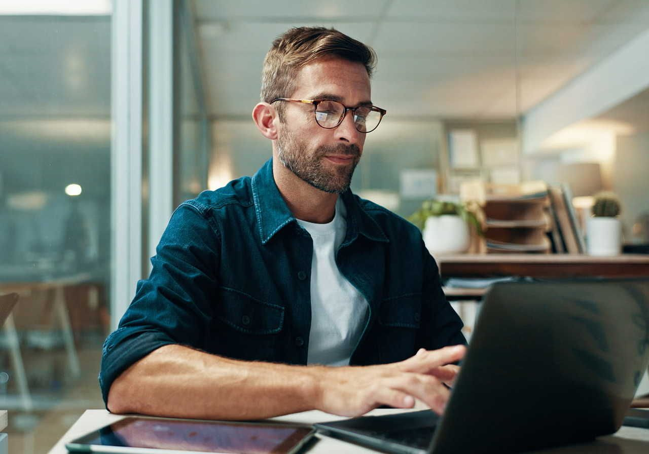A man with glasses and a beard sits in an office working on a laptop, searching for dropshipping marketing tips.