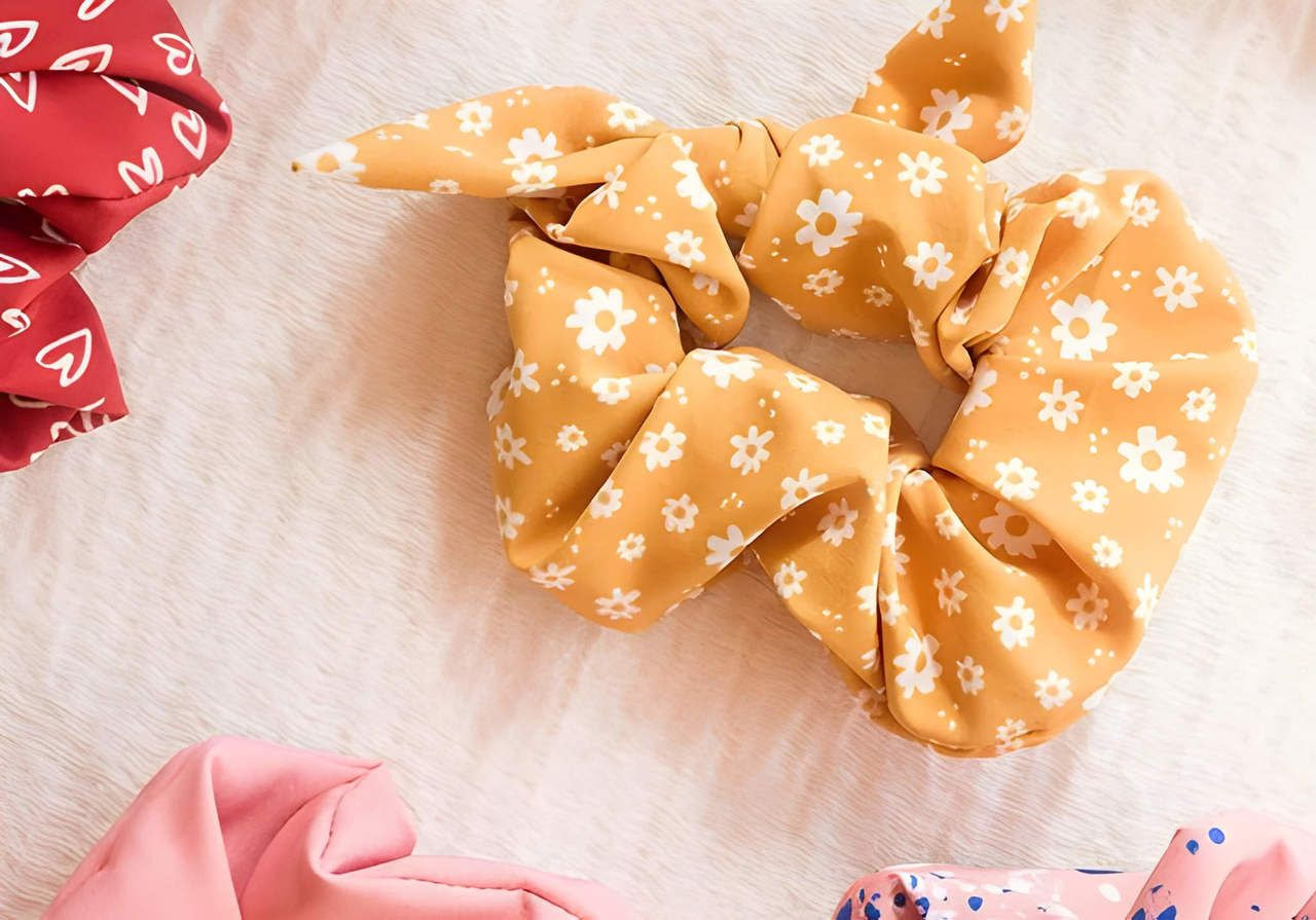 Fabric scrunchies on a textured surface. Center: a yellow scrunchie with white flowers. Surrounding it are red, pink, and speckled designs.