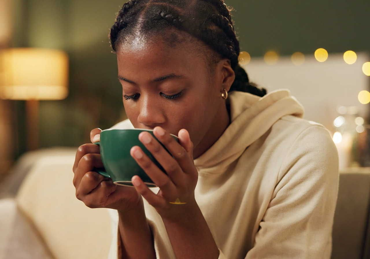 A young woman in a hoodie is sitting and sipping from a green cup, with warm lighting and a cozy background.