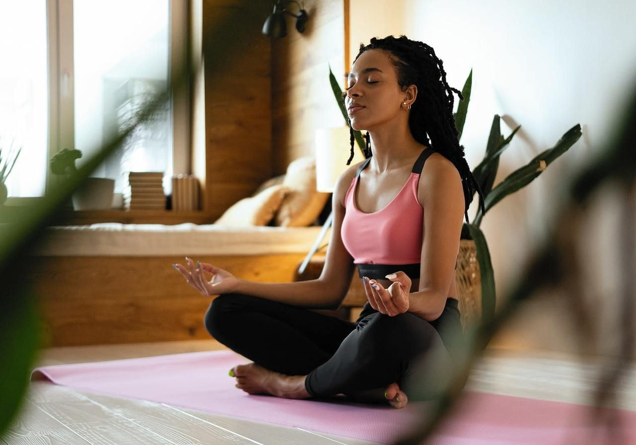 A woman sits cross-legged on a pink yoga mat, meditating peacefully with closed eyes.
