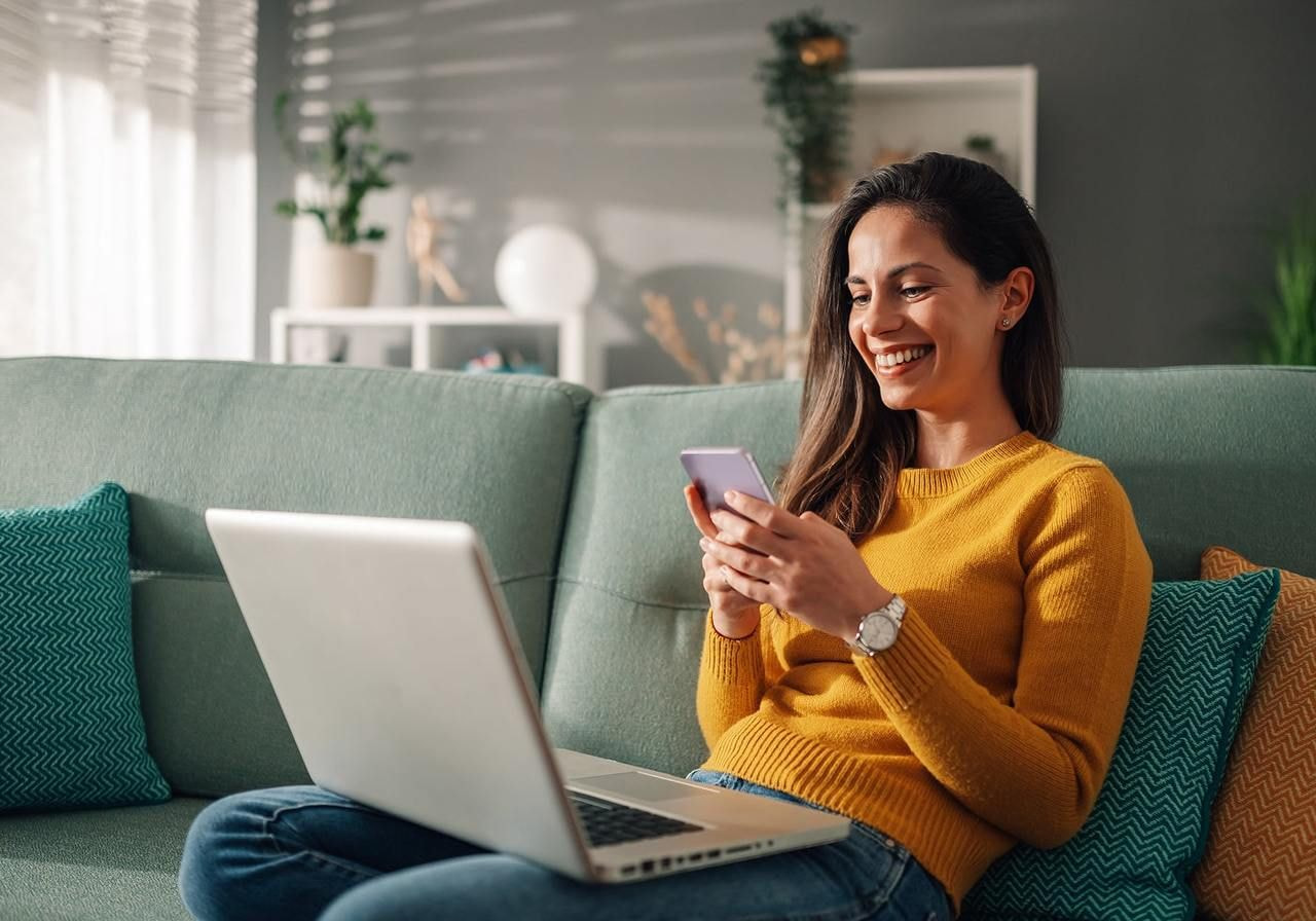 A woman in a yellow sweater sits on a green couch, with a phone in her hand next to an open laptop.
