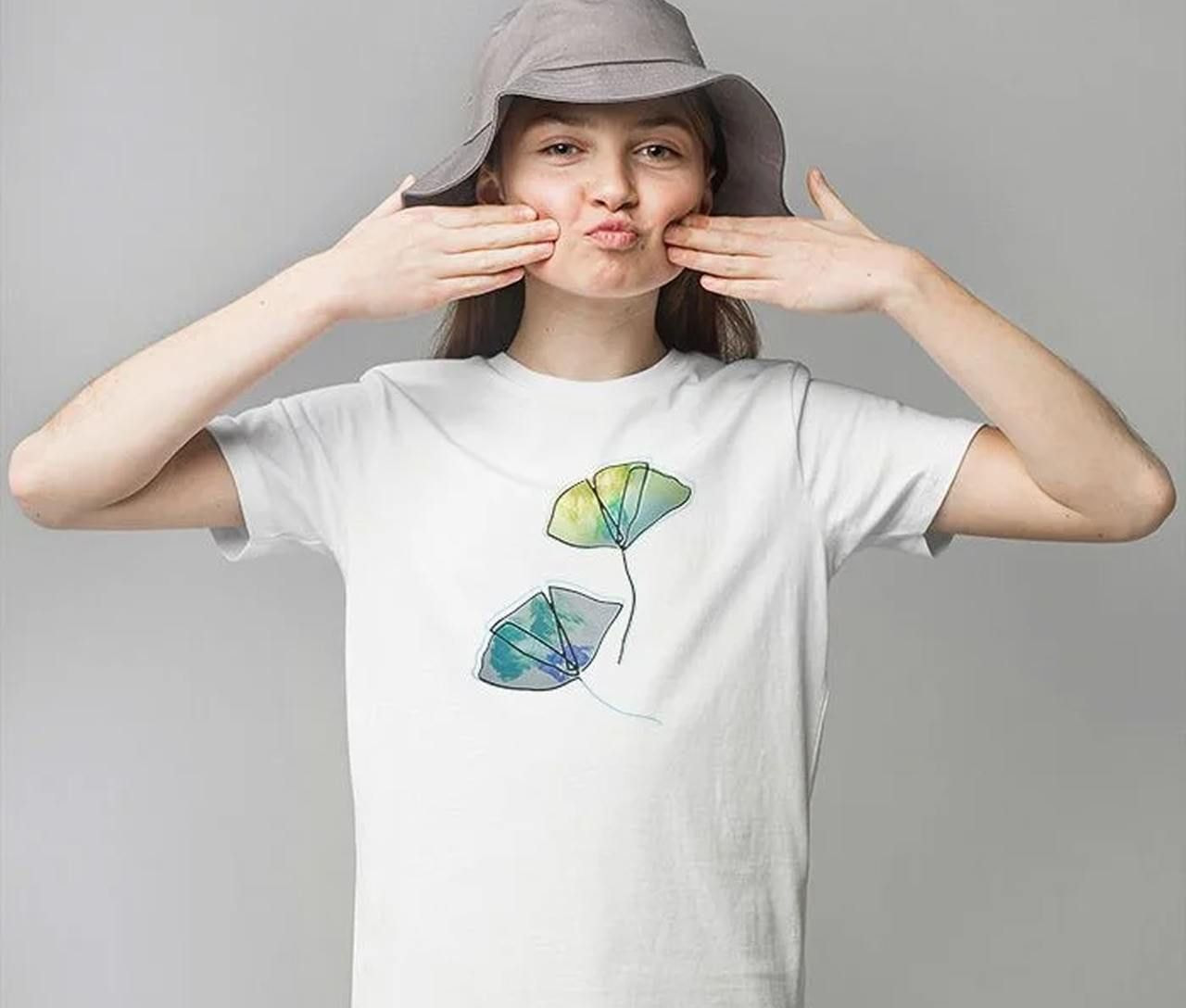 A girl wearing a white t-shirt with a green leaf design and a gray hat, with her hands raised near her cheeks.