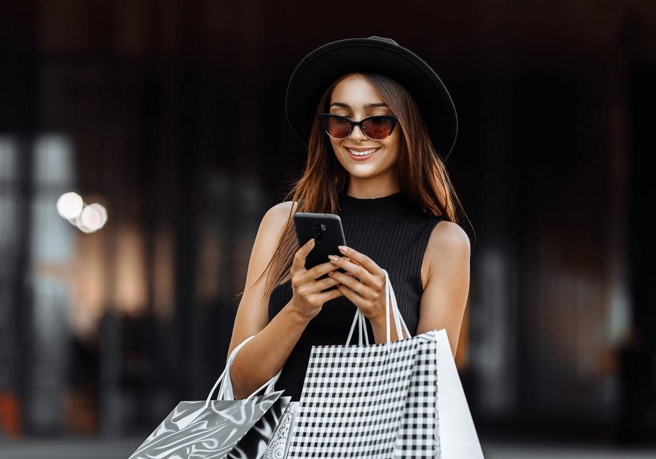 Stylish woman in sunglasses, in a black dress and hat, smiling at her phone while holding shopping bags.