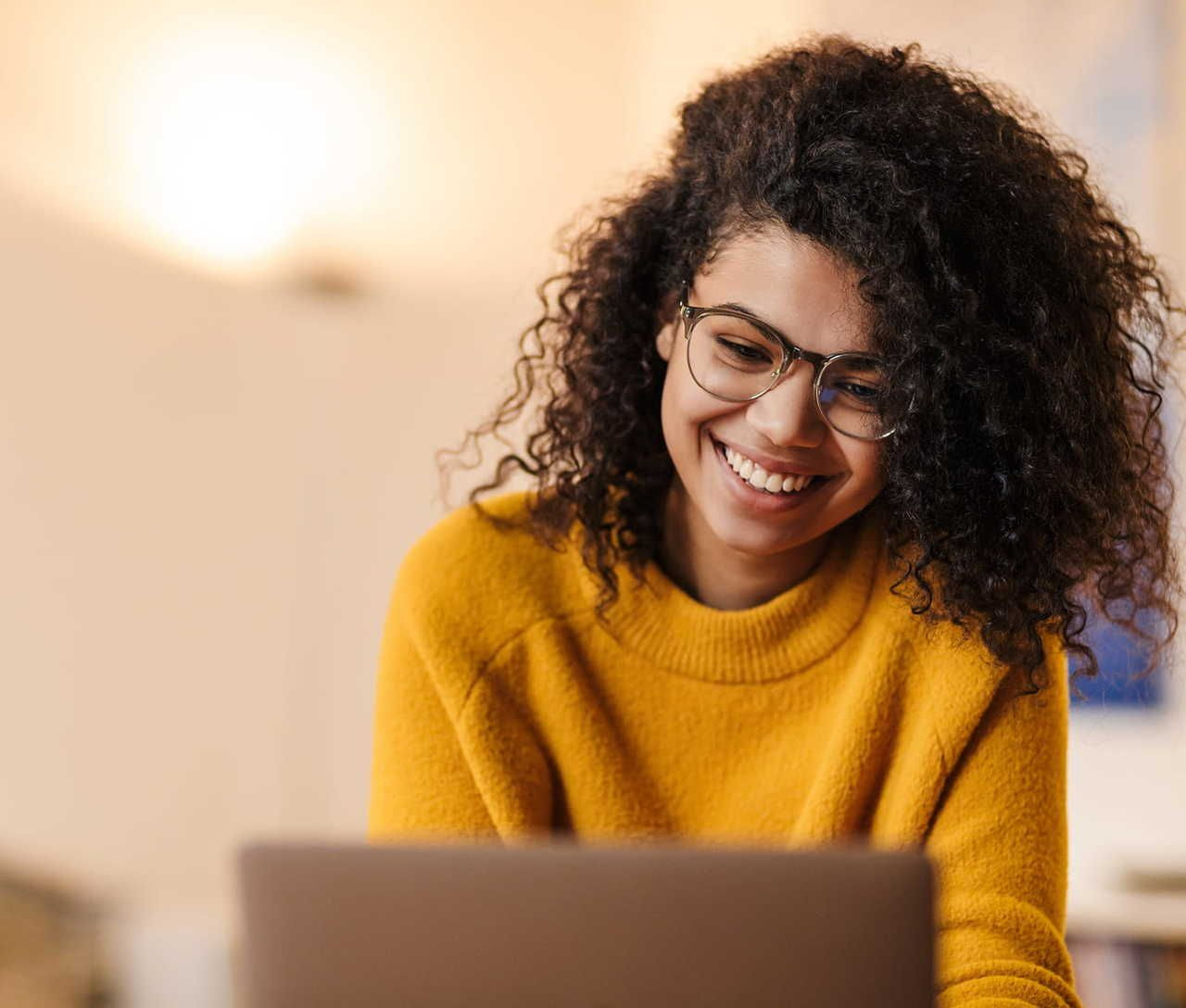 A woman with curly hair and glasses smiles while looking at a laptop. She wears a bright yellow sweater.
