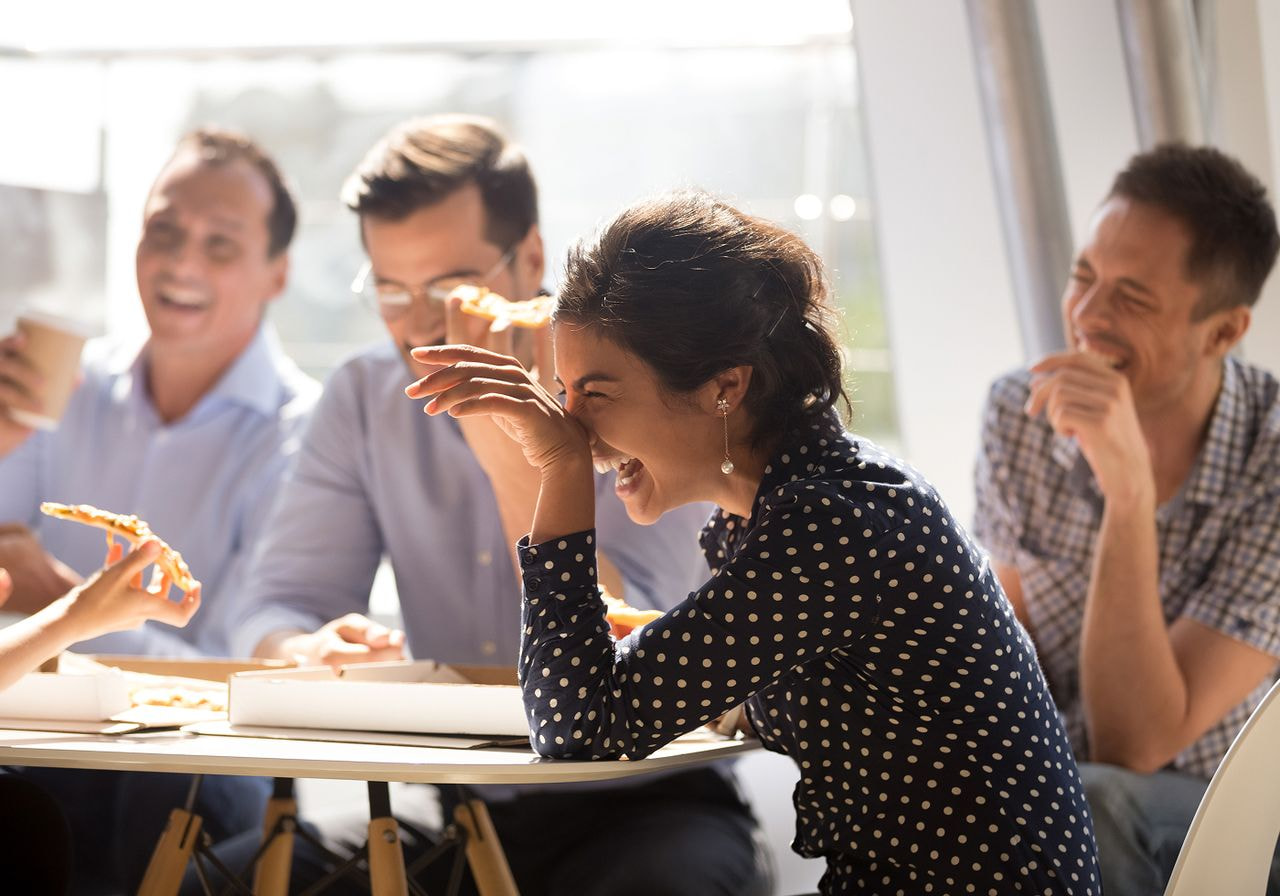 A diverse group of colleagues shares a joyful moment, laughing around a table with pizza.